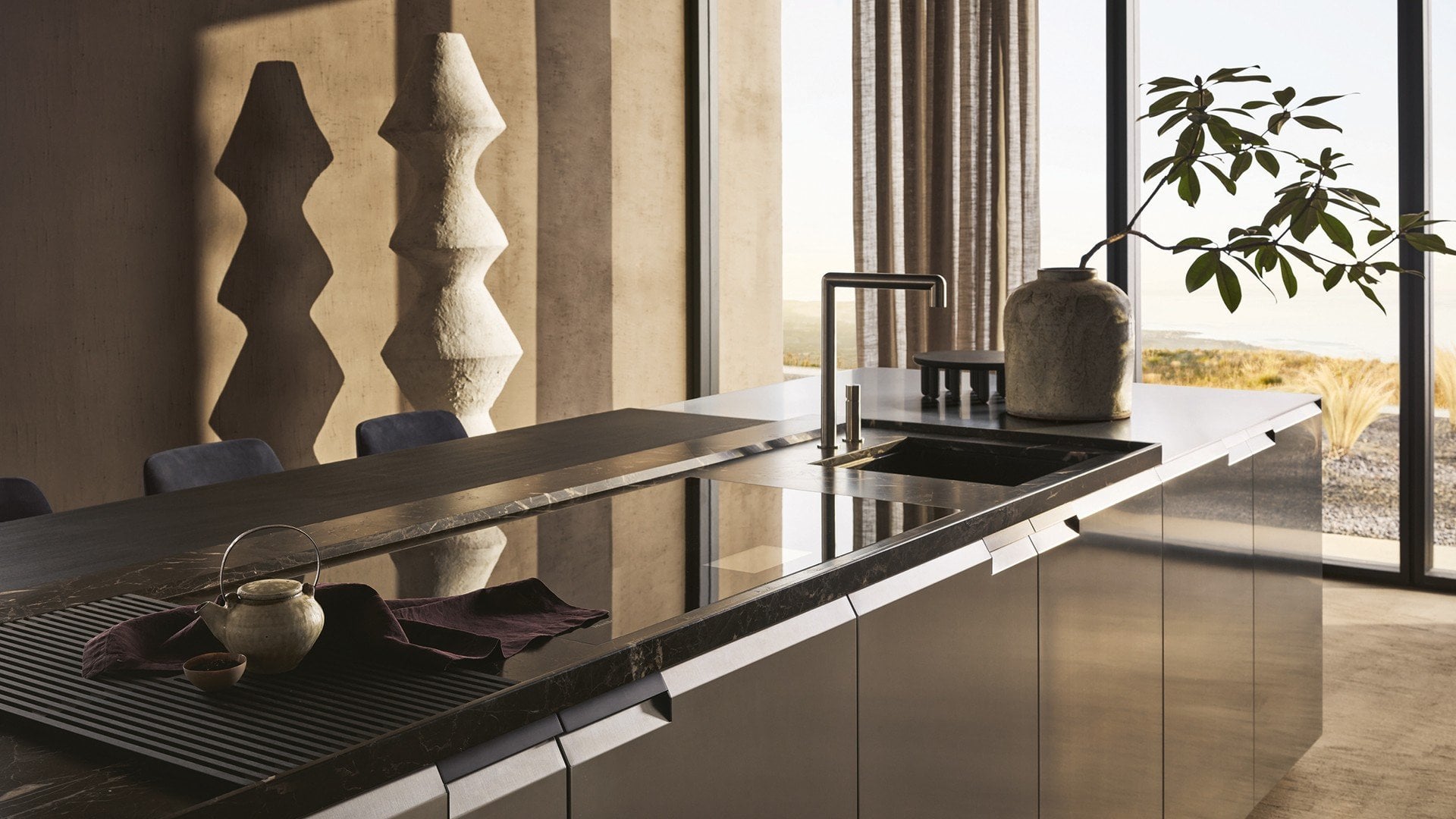 Stainless-steel island with book-matched stone edge and integrated hob, opening to a coastal view